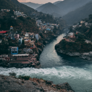 A river flows through a deep valley surrounded by steep, forested mountains. Colorful buildings are densely clustered on both sides of the river, perched on the slopes and clinging to the rocky terrain. A prominent confluence of two rivers is visible at the center, with churning white water merging into a calmer flow. The scene is framed by misty, blue-gray mountains in the background under a soft, early morning or late evening sky.