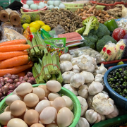 A vibrant market stall displays a variety of fresh produce, including eggs, garlic, carrots, tomatoes, okra, broccoli, and shallots, conveying abundance.