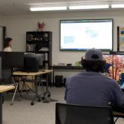 A GIS instructor leading a computer lab class in the GAM Lab, with a US map displayed on a large projector screen and students working at individual monitors.