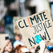 A hand-painted cardboard protest sign reading "Climate Justice Now!" with a thermometer and globe illustration, held above a crowd at a climate demonstration. Photo by Markus Spiske on Unsplash.