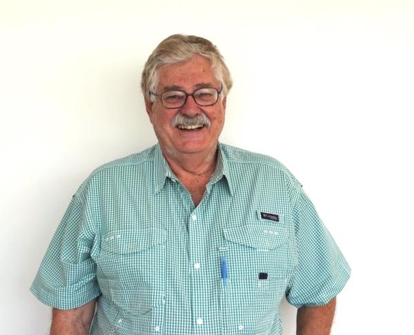 A man with graying-blonde hair and a mustache smiles. He wears a green and white checkered collared shirt and stands in front of a white background.