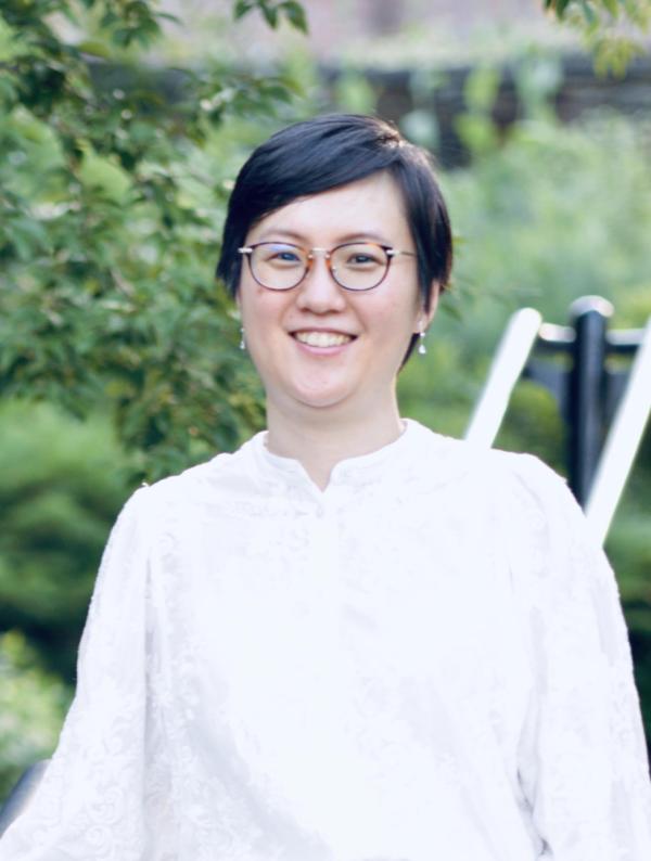 A young woman with short black hair, a white blouse, and earrings smiles in front of green foliage.