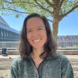 Mara has shoulder length brown hair as she stands in front of a large building