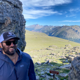 Michael on a mountain wearing a baseball cap and sunglasses smiling for the camera