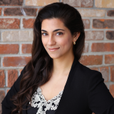 Headshot of Simone standing in front of a brick wall wearing a black blazer 