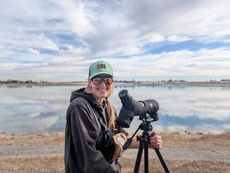Laura is standing near a lake and she has a large expensive camera on a tripod