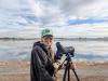 Laura is standing near a lake and she has a large expensive camera on a tripod