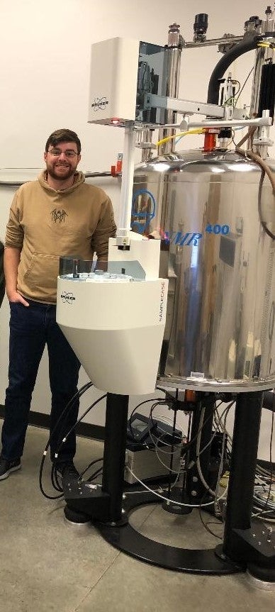 James Beham, CU Denver MS Chemistry Student, stands next to an NMR spectrometer.