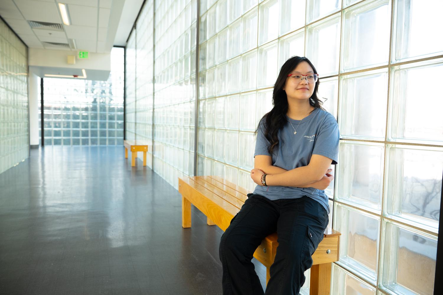 Abby sits on a wooden bench in a glass hallway, arms crossed and smiling