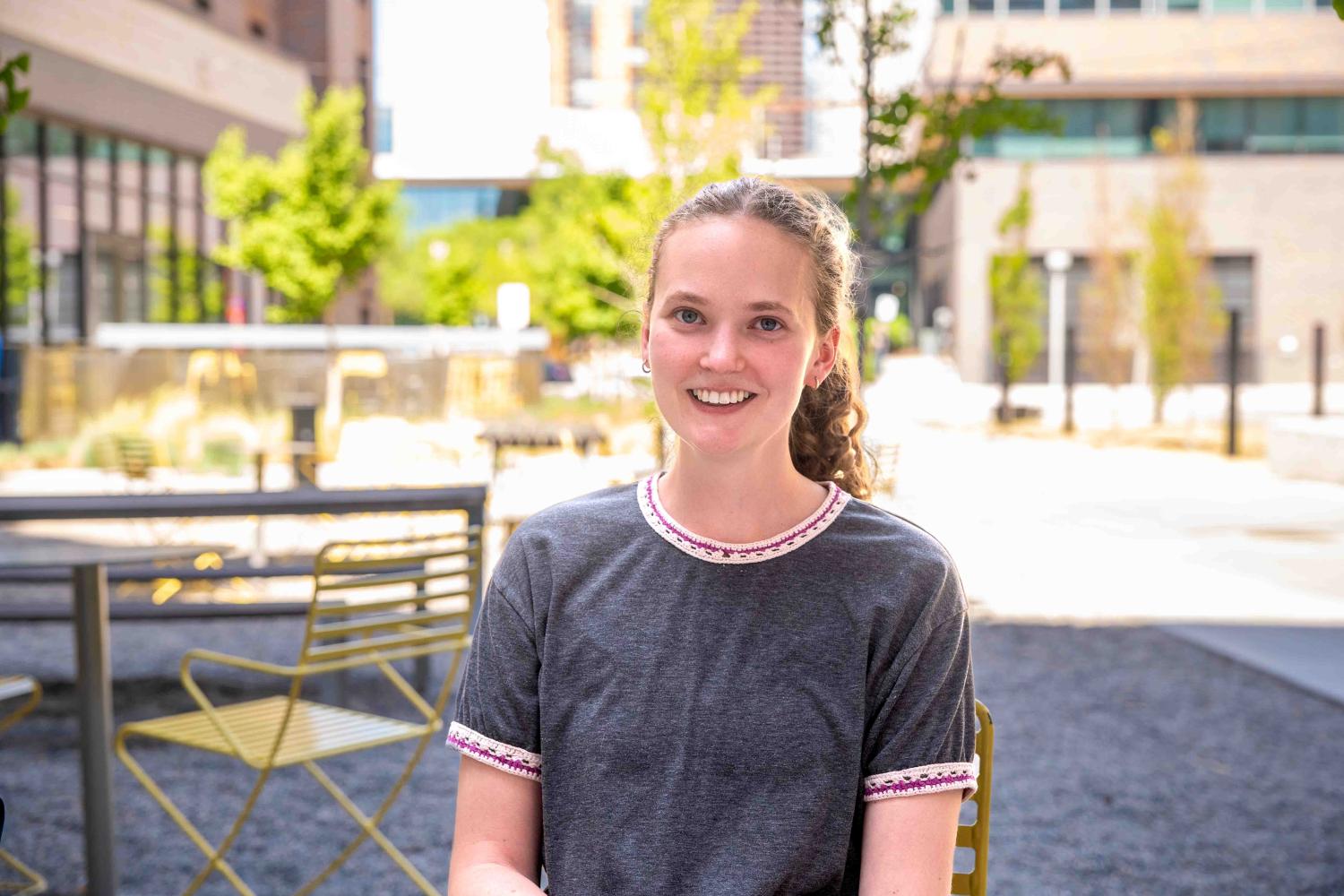 Martha smiles at the camera wearing a grey top. Behind her is a courtyard with blooming green trees and city buildings.