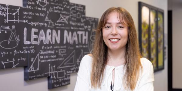 Ella stands in front of a chalkboard that reads Learn, wearing a white top.