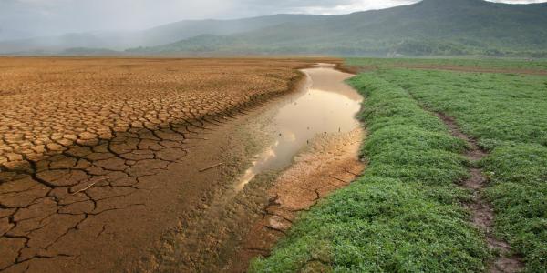 A landscape showing a wide stretch of cracked, dry earth beside a narrow ribbon of muddy water, with green vegetation on one side. Mountains and heavy clouds loom in the background, suggesting drought conditions in a once-wet area.