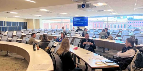 n interior view of a wide, sunlit classroom with tiered, curved wooden desks and grey ergonomic chairs. Participants are gathered in small clusters talking, while a blue presentation screen in the background displays discussion prompts titled "Layer 1: What Went Wrong, and When?" Large windows offer a view of downtown Denver buildings.