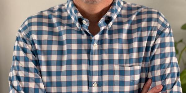 Hani Mansour smiles at camera. He is wearing a blue and white plaid button up shirt, and green rimmed glasses.