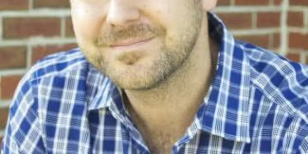 Wayne Miller, with short light-brown hair and a trimmed beard, is sitting in front of a brick wall. He is wearing a blue plaid shirt and looking at the camera with a slight smile