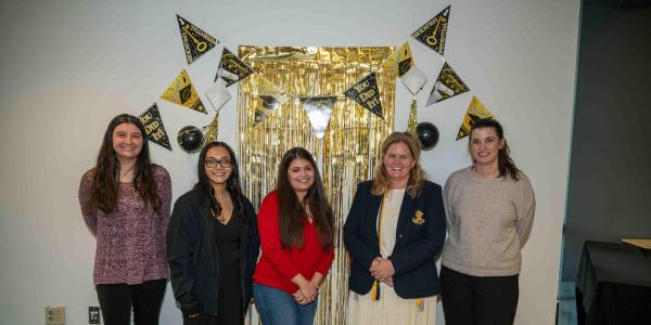 Five CLAS Outstanding Graduate Award recipients stand together in front of a gold graduation-themed backdrop, smiling during the Fall 2025 recognition event.