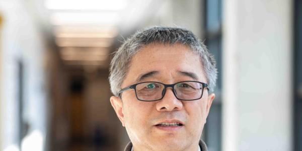  Dr. Haobin Wang, Professor of Chemistry at the University of Colorado Denver, poses in a bright university corridor. He is wearing glasses and a gray plaid polo shirt, with a softly blurred hallway visible behind him