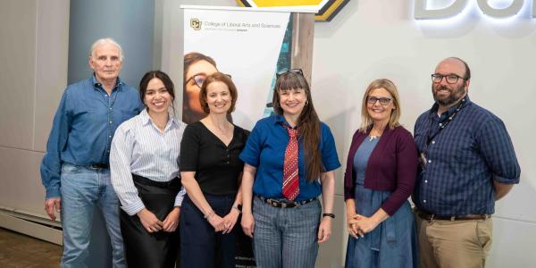 From left to right Jim Grigsby, Vanessa Navarro-Rodríguez, Anna Warrener, Nicky Beer, Lisa Keranen, and Michael Moore. They stand in front of a University of Colorado Denver sign and a banner for the College of Liberal Arts and Sciences 