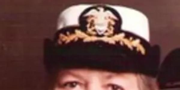 Professor Mary Rose Sullivan in a formal U.S. Navy uniform, wearing a white cap with gold trim and insignia, smiling slightly against a dark background.