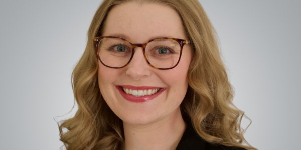 Kayley smiles at at camera with glasses and a black blazer on.
