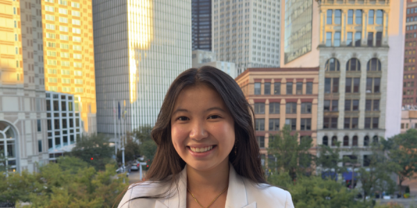 Kim stands smiling at camera on a rooftop. She is wearing a white blazer.