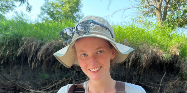 Martha smiles at camera in a riverbed, carrying a fish. She is in beige outdoor gear.