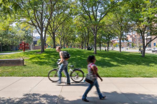 Pedestrians and a cyclist pass a tree-filled campus green on a sunny day.