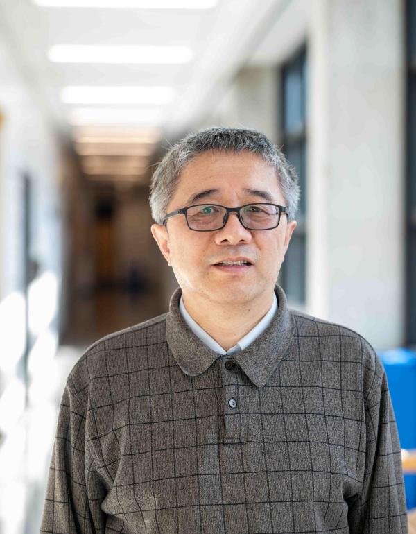 Dr. Haobin Wang, Professor of Chemistry at the University of Colorado Denver, poses in a bright university corridor. He is wearing glasses and a gray plaid polo shirt, with a softly blurred hallway visible behind him