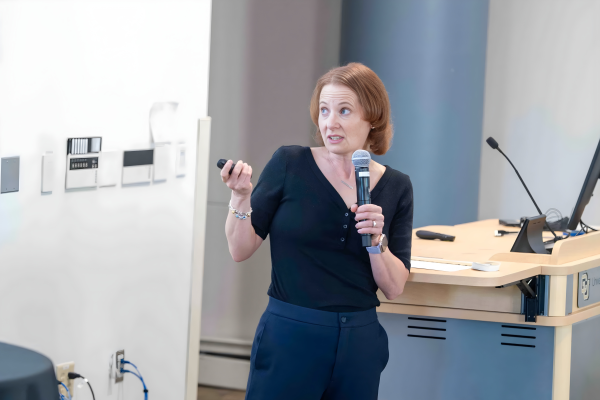 Anna Warrener speaks into a handheld microphone while gesturing with a remote during a presentation, standing beside a podium.