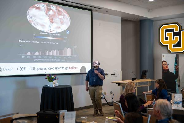 Michael Moore gives a presentation in a conference room, holding a microphone and looking up at a large screen displaying a global temperature map and climate data charts. Audience members sit at tables in the foreground, and University of Colorado Denver signage and a College of Liberal Arts and Sciences banner are visible in the room.