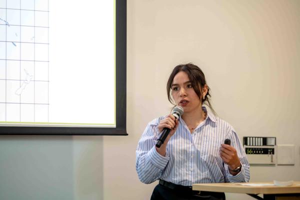 Vanessa Navarro-Rodríguez speaks into a handheld microphone while giving a presentation, standing beside a projected slide and holding a remote, dressed in a blue striped shirt.