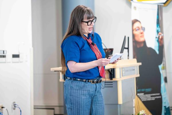 Nicky Beer wearing glasses and a blue shirt with a red tie reads from note cards while speaking at a podium.