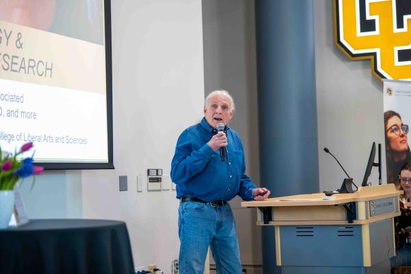 Jim Grigsby in a blue shirt speaks into a handheld microphone while presenting beside a podium.