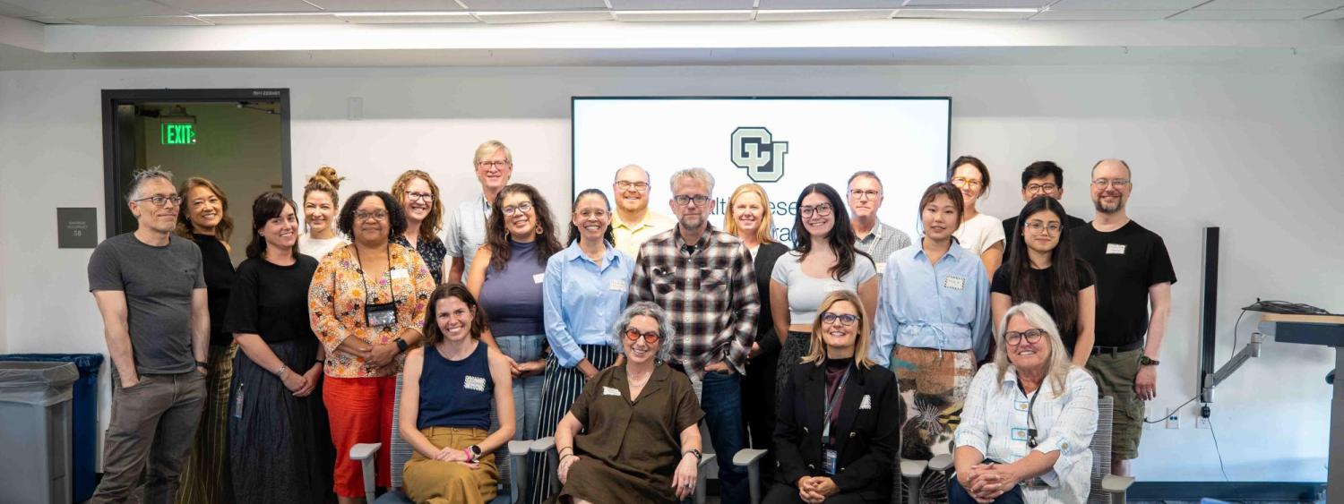 Group photo of the CU Denver Health Research Collaborative kickoff, with approximately two dozen participants gathered in a conference room, standing and seated in front of a screen displaying the CU Denver logo.