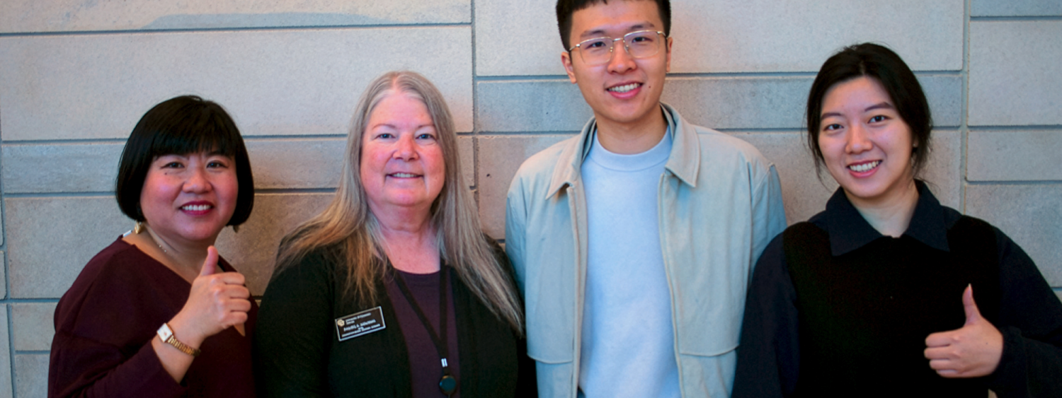 a woman in a burgundy top, a woman with long gray hair wearing a black cardigan and CU Denver badge, a man in glasses and a light jacket, and a woman in a dark dress. All appear friendly and posed for a group photo at the Eleventh Scholars Summit event.