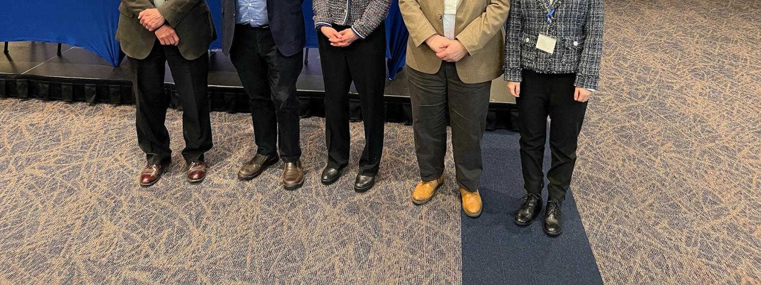 Gemini said A group of five professionals stands in a conference room in front of a large screen displaying details for a "Generative AI Frontier" event in 2026. The group consists of three men and two women, dressed in business casual and professional attire, all wearing blue lanyards with name tags. The man second from the left is David Hildebrand. He is wearing a navy blue blazer over a light blue button-down shirt and dark trousers, smiling at the camera.
