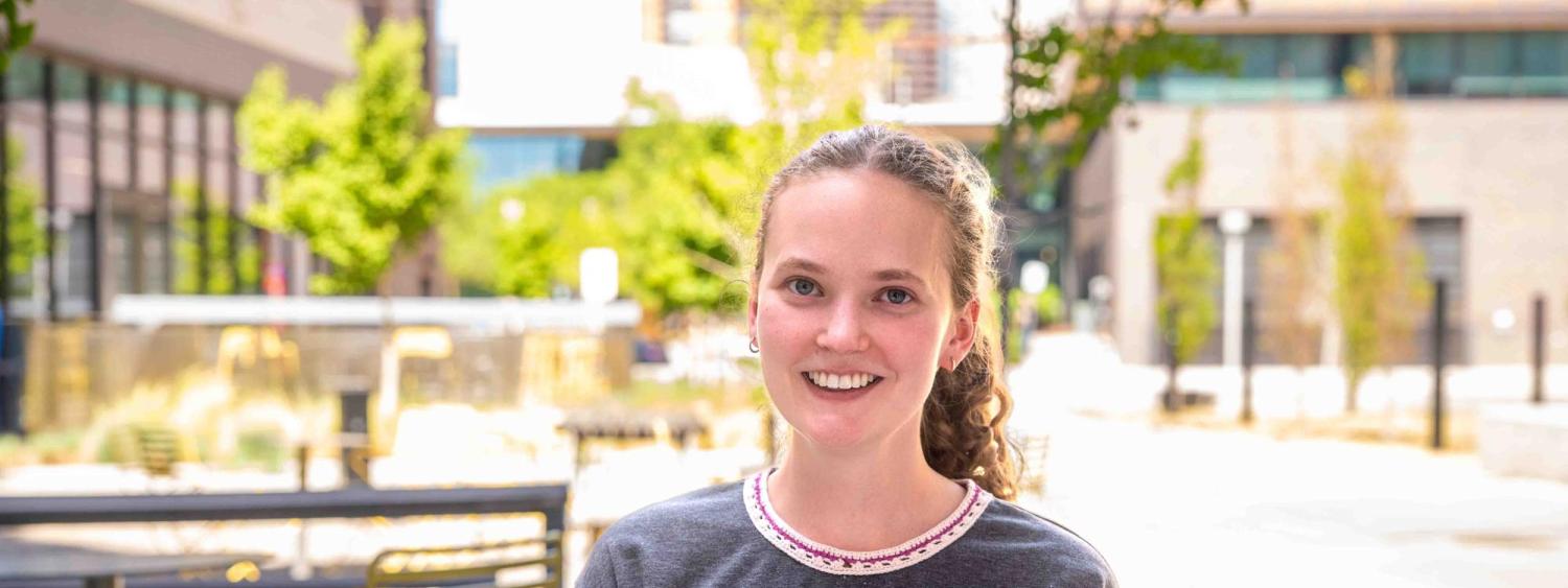 Martha smiles at the camera, wearing a grey tshirt. The background is a couryard with blooming green trees and city building.