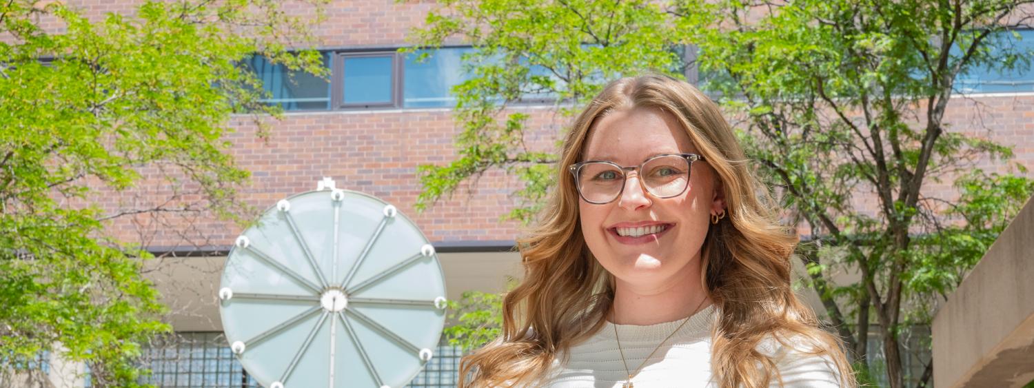 Kayley stands smiling with arms crossed in front of clock