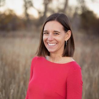 Liz D. standing outdoors in a field of tall grass, wearing a bright red three-quarter sleeve dress. She has straight, shoulder-length brown hair and is facing the camera, with trees softly blurred in the background and warm natural light.