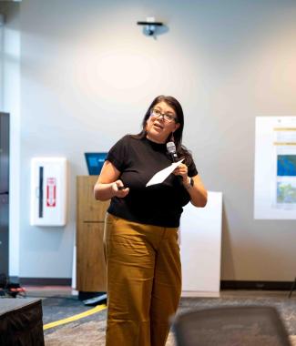 Edelina Burciage gives a presentation at CLAS Mixtape. She is wearing yellow pants, a black top, and glasses.