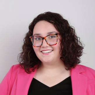 Kayla Ahr smiles at camera, she is wearing a pink blazer and glasses against a white backdrop.