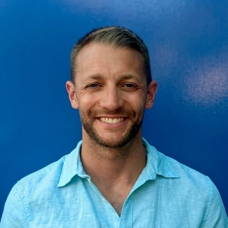 Cameron smiles at camera in a blue t-shirt against a blue wall.