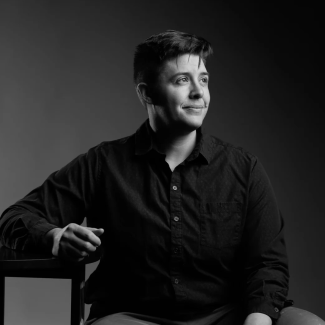 A black and white photo of Rachel where she sits on a chair, looking to the left, in black shirt.