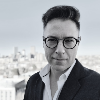 A black and white photo of Adam wearing a black blazer, in framed glasses. 