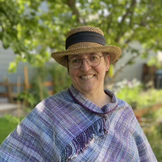Shana Médah smiles with a tweed hat, outside, in a blue and purple top. 