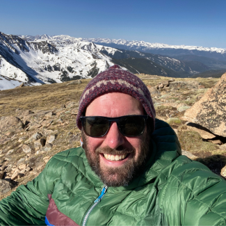 Michael Moore stands at a mountain top, wearing a burgundy beanie, a green jacket, and sunglasses.
