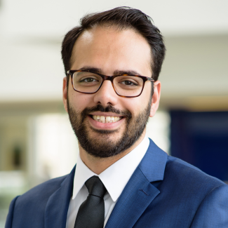 Farhad smiles at camera, wearing glasses and a suit in a office like setting.