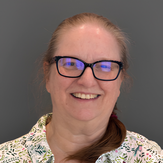 Catherine smiles to camera, against a gray backdrop. She is wearing a floral button up and glasses.