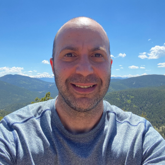 Adam Spiegler smiles at camera, outdoors wearing a grey tshirt amongst the mountains.