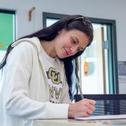 A student wearing CU Denver gear writing at a counter inside a brightly lit campus building.
